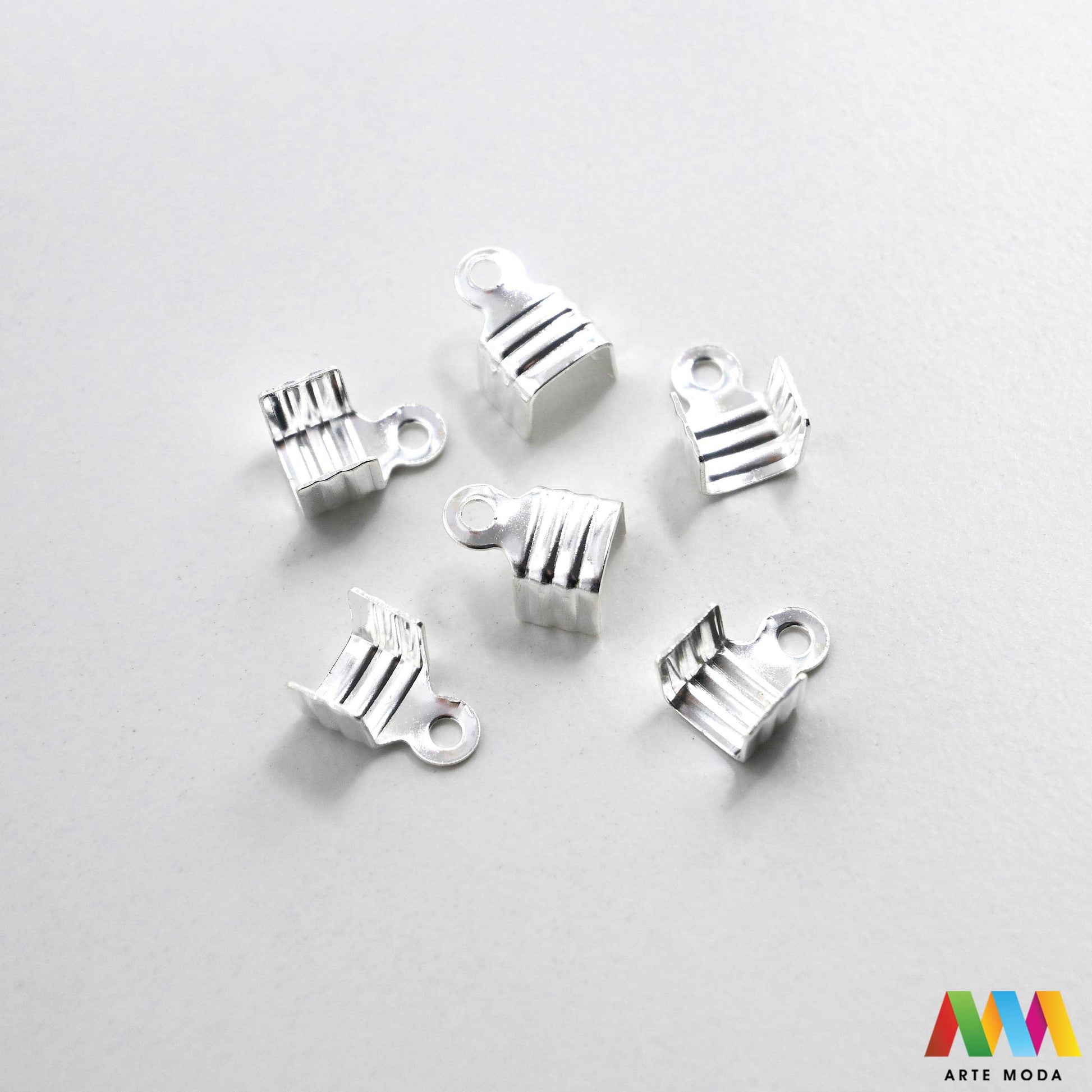 Metalni stiskači i završeci sa zubima za izradu nakita u srebrnoj, zlatnoj i brončanoj boji složeni na radnoj površini. Prikazani su ravni stiskači za tekstilne trake, baršun, antilop i plosnate kožice s integriranom omčicom za spajanje s kopčama i produžnim lančićima. Kvalitetan repromaterijal za uredno fiksiranje krajeva pri izradi choker ogrlica, narukvica i modnih dodataka u DIY radionicama za ručnu izradu unikatnog nakita s nazubljenim rubovima koji jamče čvrst prihvat bez ispadanja materijala.